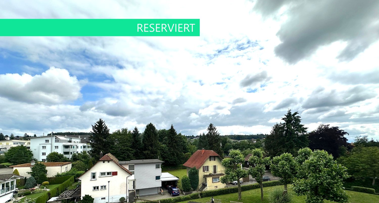 Several houses, greenery, cars parked in front, distant buildings in the background, cloudy sky