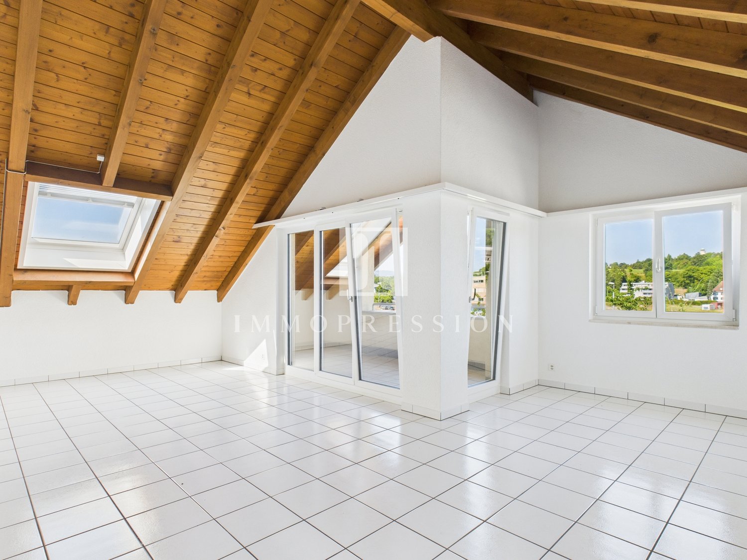 Spacious attic apartment with wooden beamed ceiling, large windows, and sliding glass doors leading to a balcony or terrace. The room has a tiled floor and an open layout.