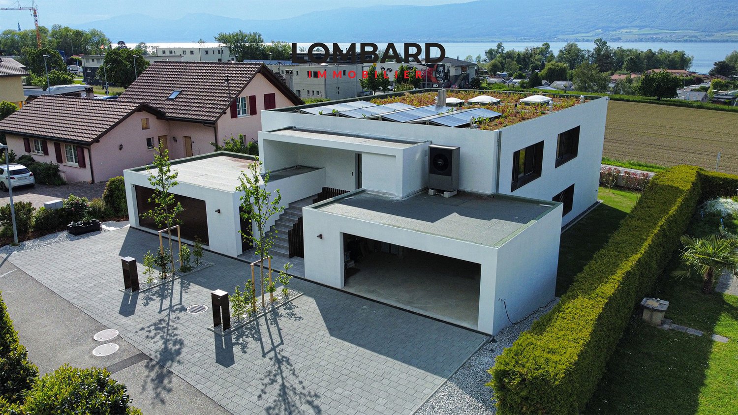 Modern white house with a green roof, solar panels, garage, and landscaped garden with hedges. It is a two-story property with a view of the mountains and a lake.