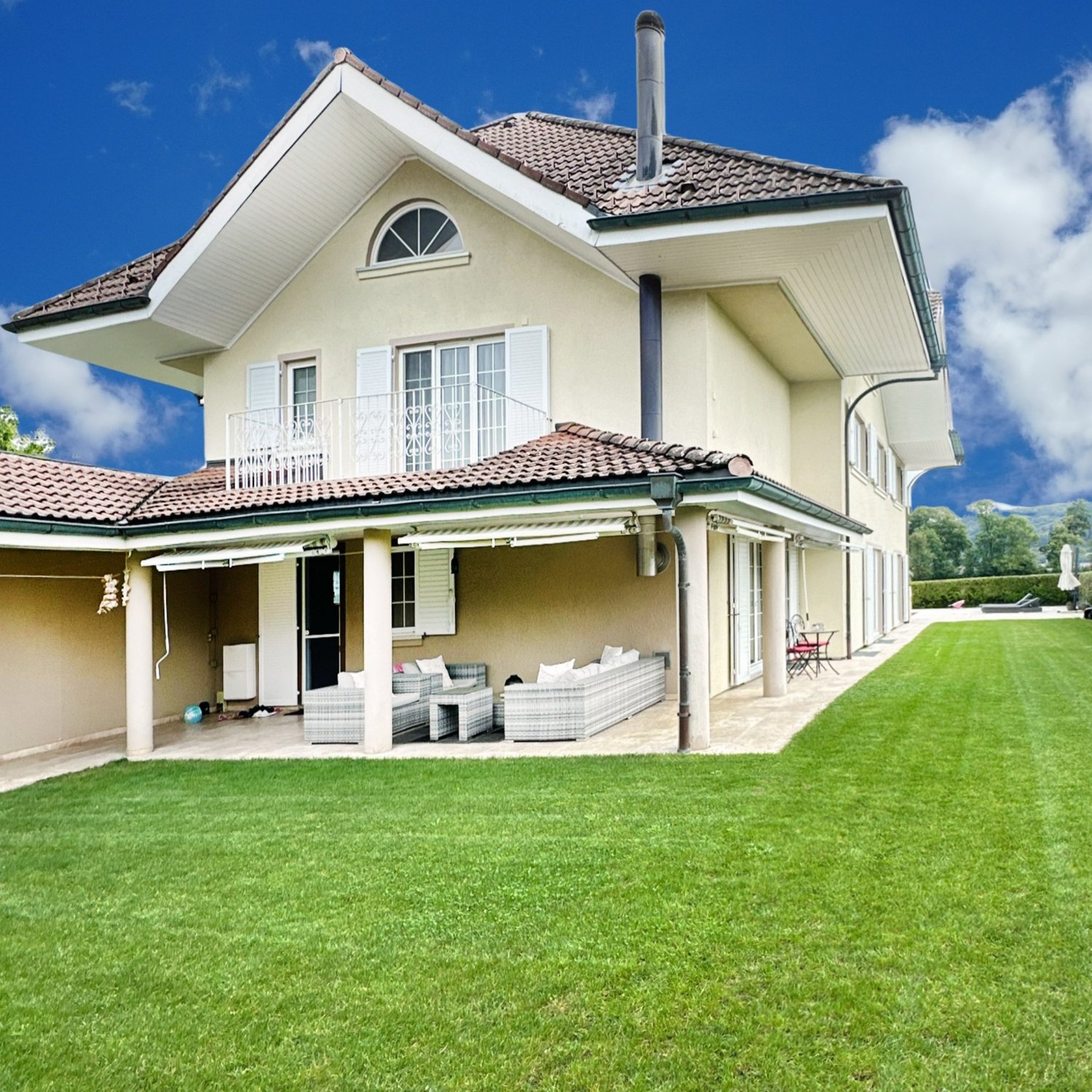 Two-story villa, beige exterior, red roof, large windows, covered porch, garden with lawn, lounge chairs, outdoor seating, balcony with railing, expansive view of greenery and mountains in the background