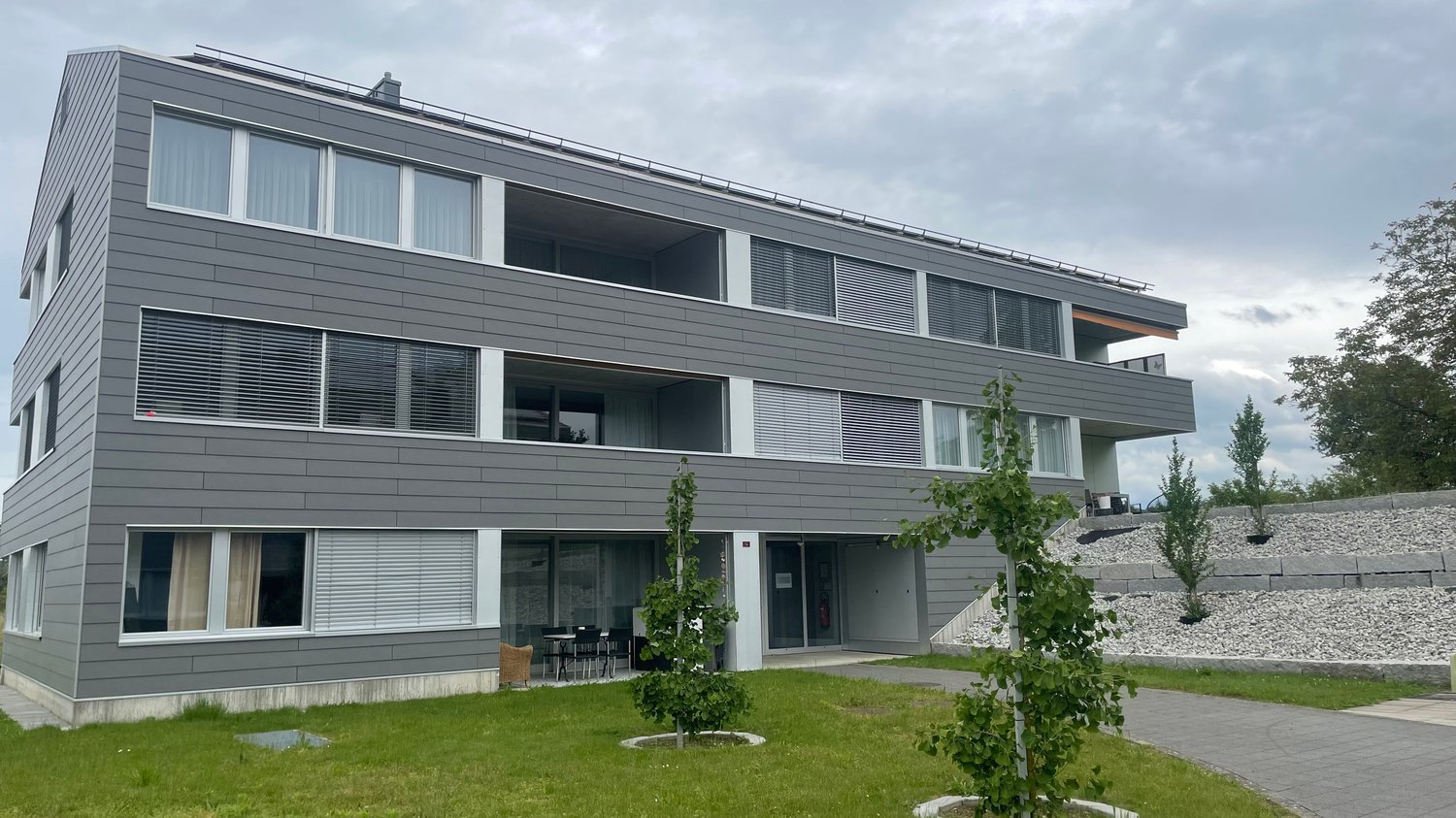 Modern gray apartment building with multiple floors, balcony on the second floor, glass windows, gray exterior, green lawn, gravel path, small plants, patio furniture in front.