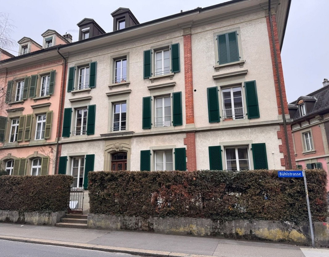 4-storey apartment building, multiple windows with green shutters, central entrance with a small garden, cobblestone street, adjacent buildings