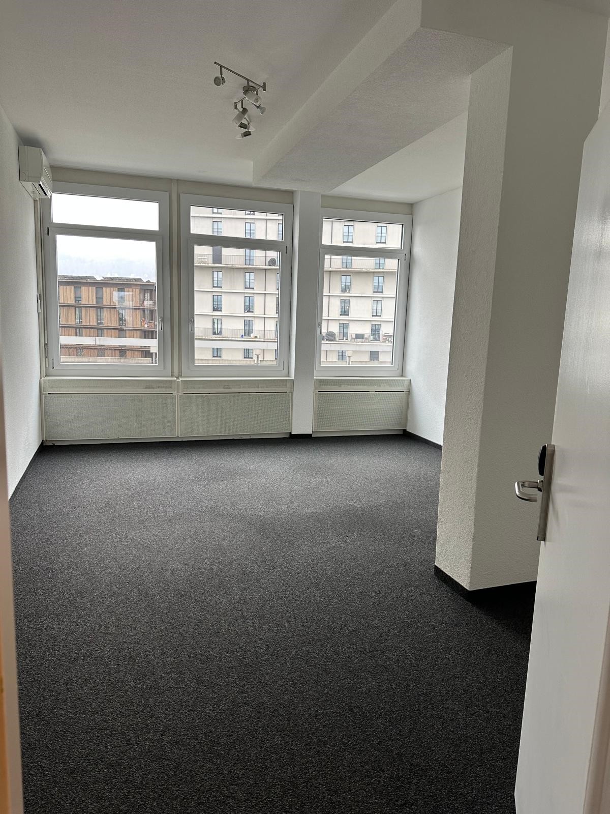 This image shows an empty room with large windows overlooking a city skyline. The room has white walls, a grey carpeted floor, and a track lighting fixture on the ceiling. The room appears to be a living or office space.