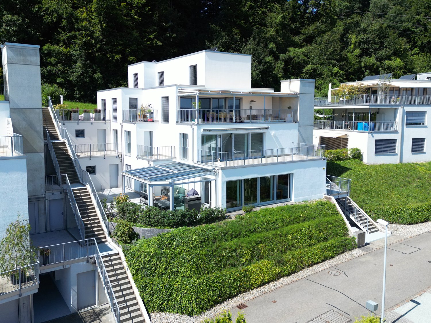 Multi-story residential complex, modern white architecture, multiple balconies, large windows, central greenery and shrubbery, steep stairways, surrounded by trees
