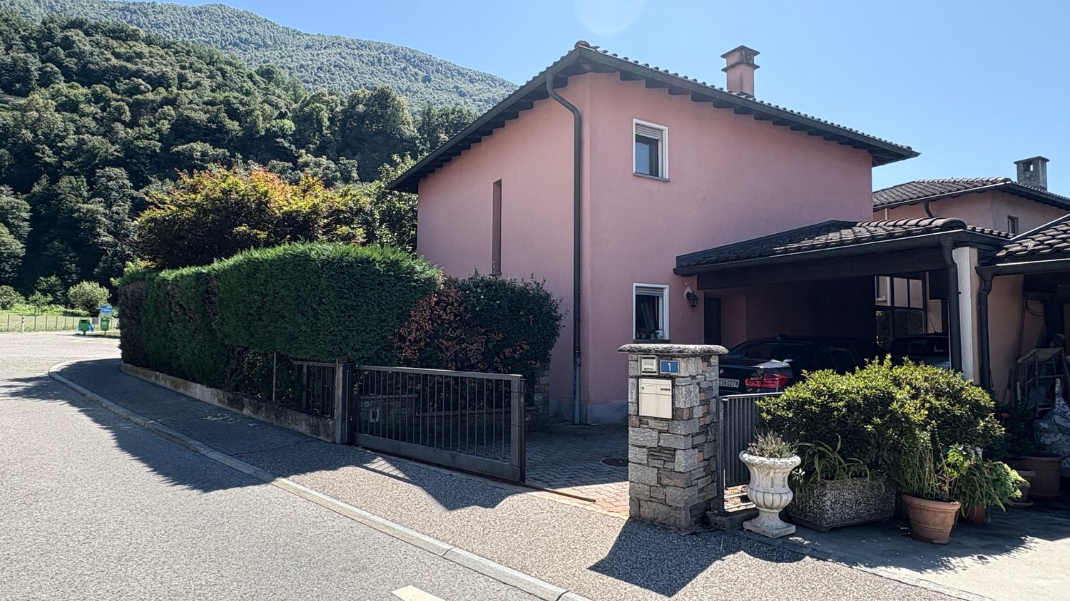 Two-story pink house with a tiled roof, surrounded by trees and greenery. The house has a garage and a stone wall with a gate in the front. There are potted plants and other decorative elements around the entrance.