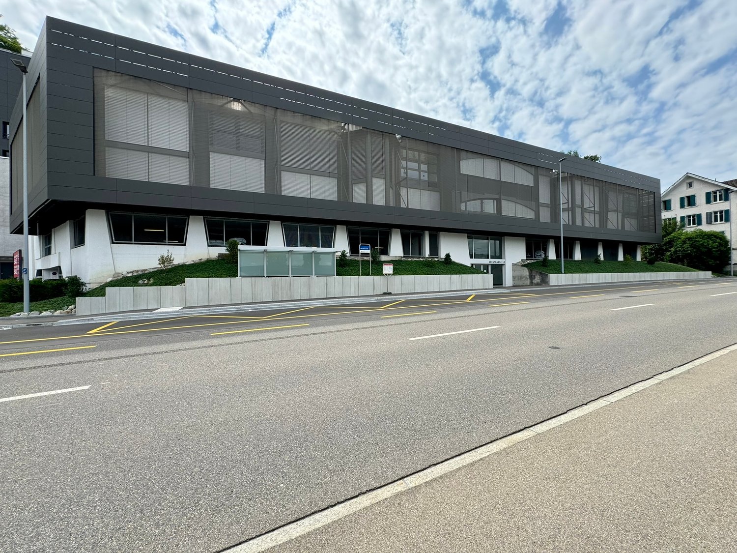 A large modern commercial or office building with a sleek, gray exterior and large windows. The building has a curved roofline and is situated next to a road with yellow lane markings.