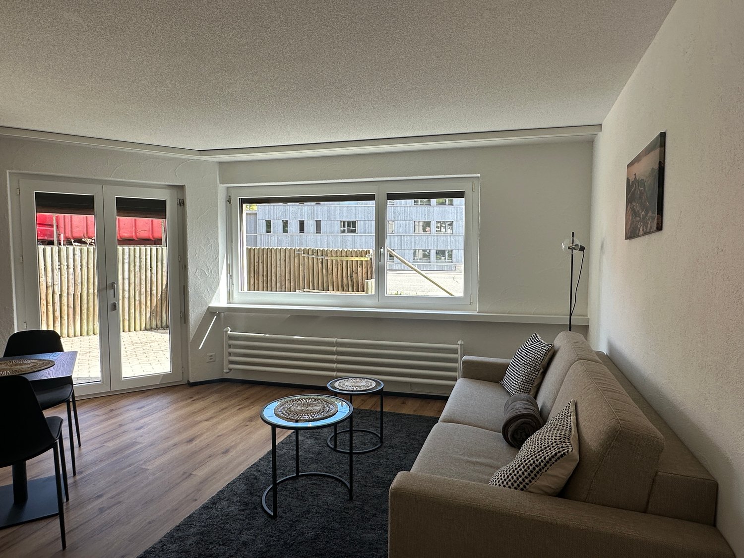 A modern living room with wooden flooring and neutral walls, featuring a beige couch with pillows, two small round tables, and sliding doors leading to an outdoor area. The room is well-lit by a large window facing the outside.