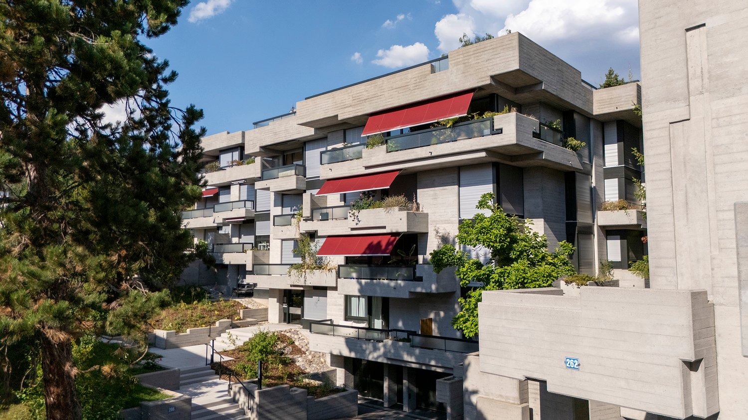 Modern architectural design, concrete building, multiple balconies with red awnings, green trees, grass, and shrubs, pathways with steps