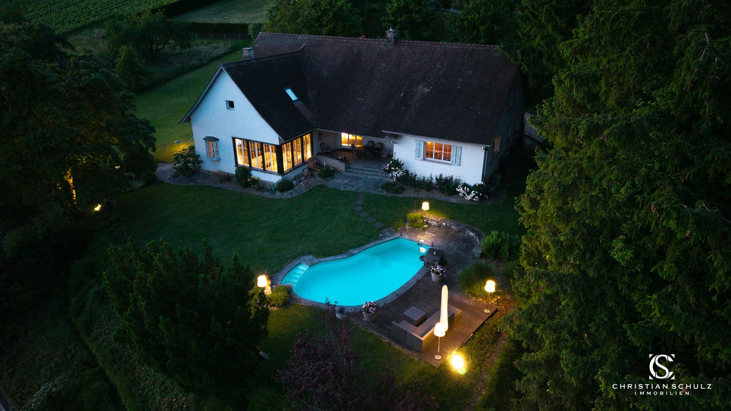 White house, brown roof, pool, deck, surrounded by trees, multiple outdoor lights