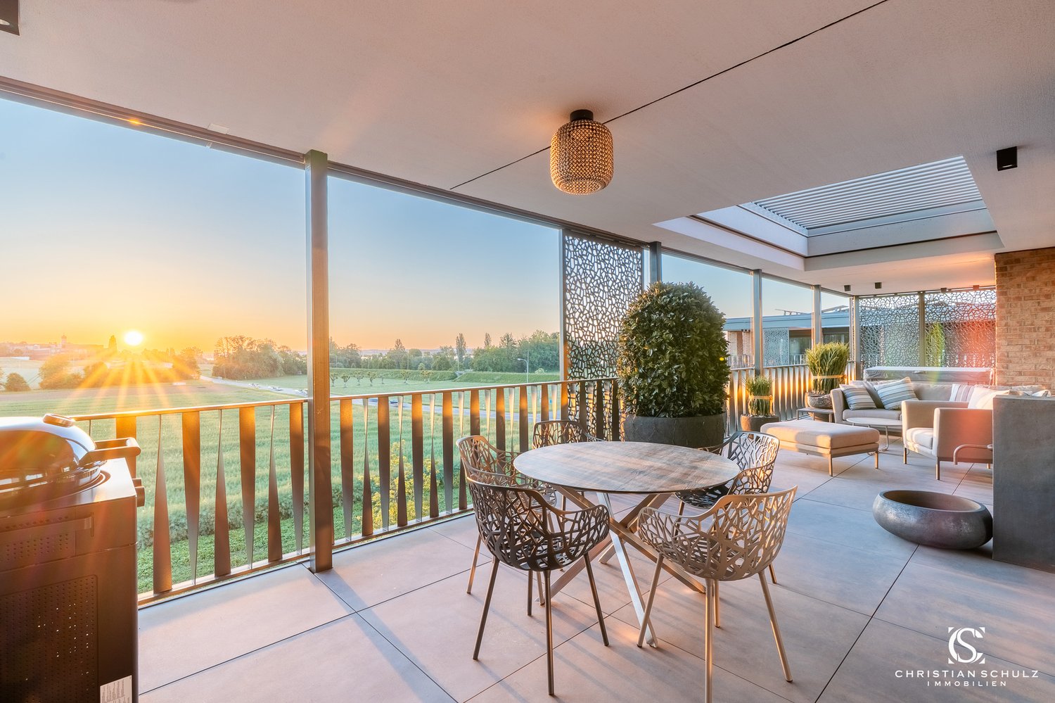 Outdoor balcony, glass walls, table, chairs, greenery, round bowl, metal griller, metal ceiling lamp, metal railings