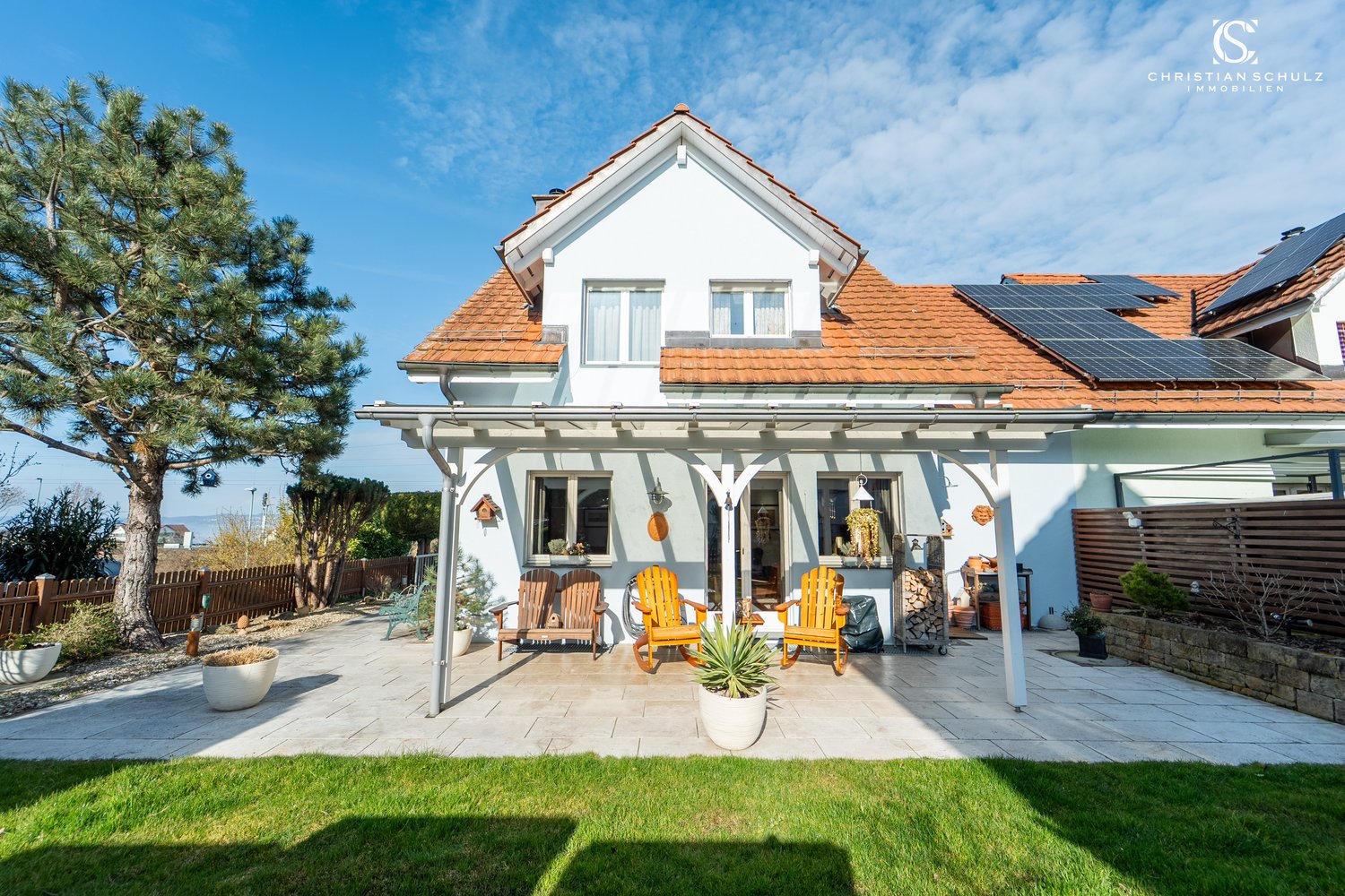 white house, red roof, solar panels, patio with wooden chairs, potted plants, wooden fence, lawn