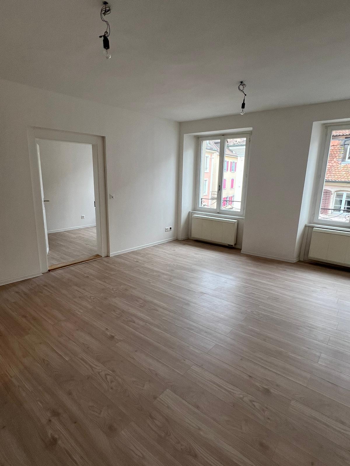 Empty room with hardwood floors, large windows, white walls, a door leading to another room.