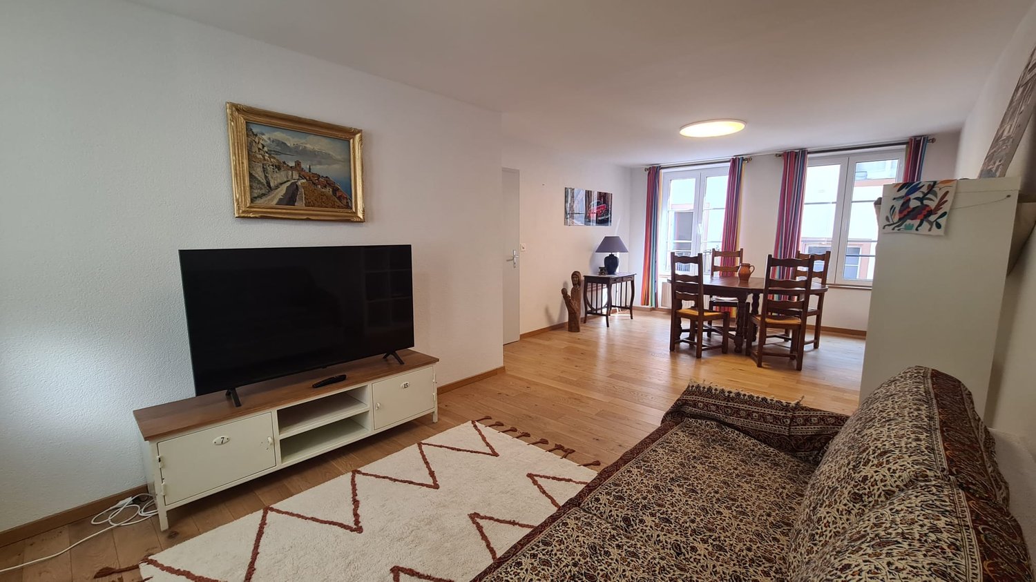 Spacious living room with hardwood floors, white walls, large windows, a TV stand, a sofa, a dining table with chairs, and artwork on the walls.