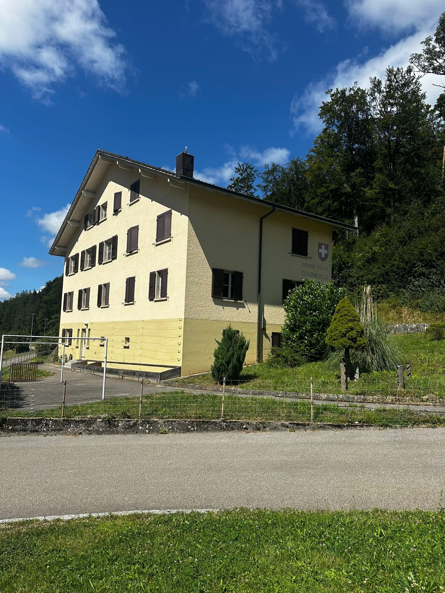 A multi-story yellow building with a Swiss flag on the roof, surrounded by trees and a grassy area.