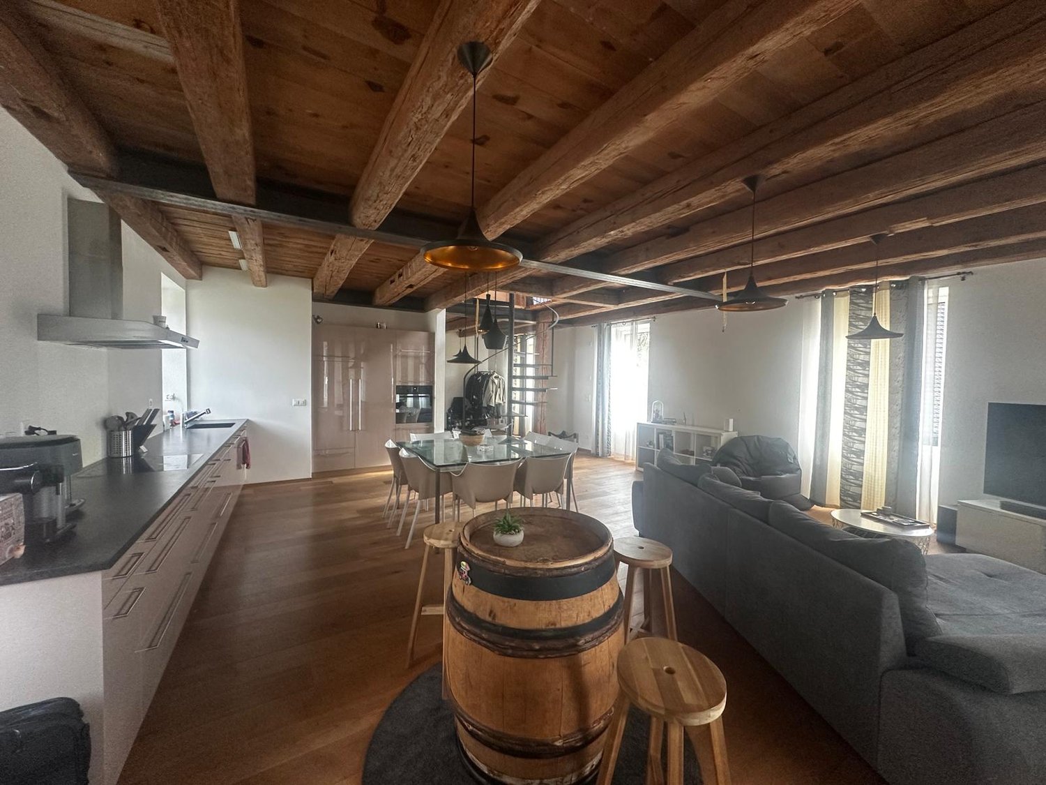 Wooden beams, modern kitchen, glass dining table, dining chairs, grey sofa, black carpet, barrel bar stool, wooden lamps