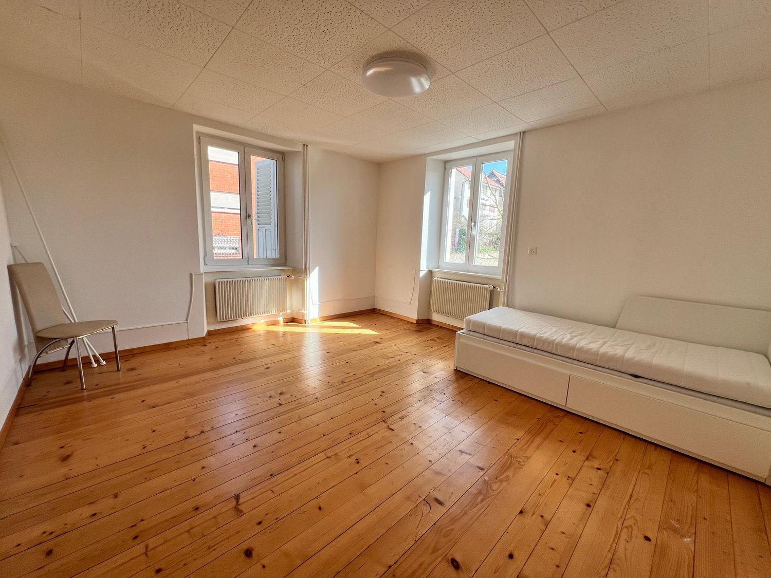spacious room, wooden floor, 2 windows, white walls, simple furniture