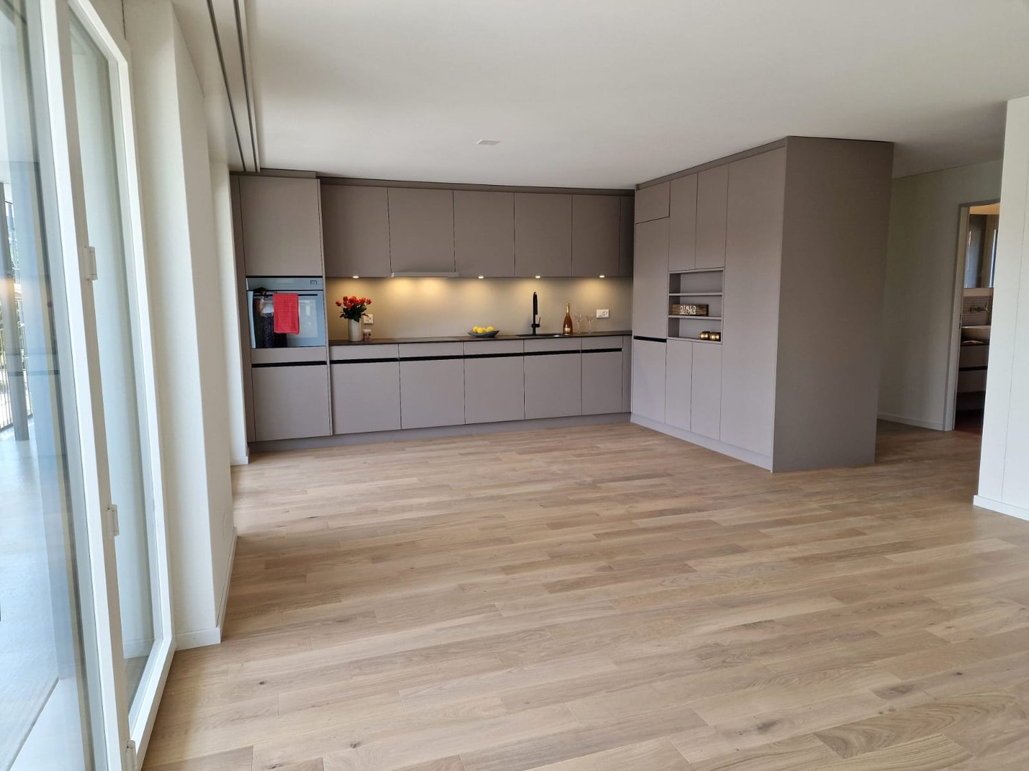 Kitchen with gray cabinetry, wooden flooring, integrated appliances, window, vase with flowers, and sink.