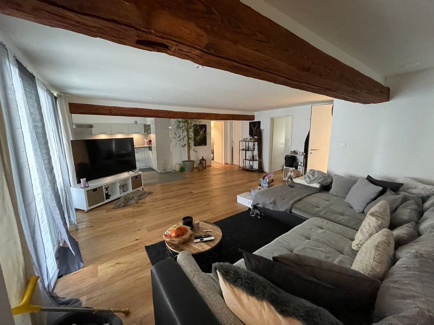 Living room with hardwood floor, large sofa, TV, wooden beams, white curtains, wooden coffee table