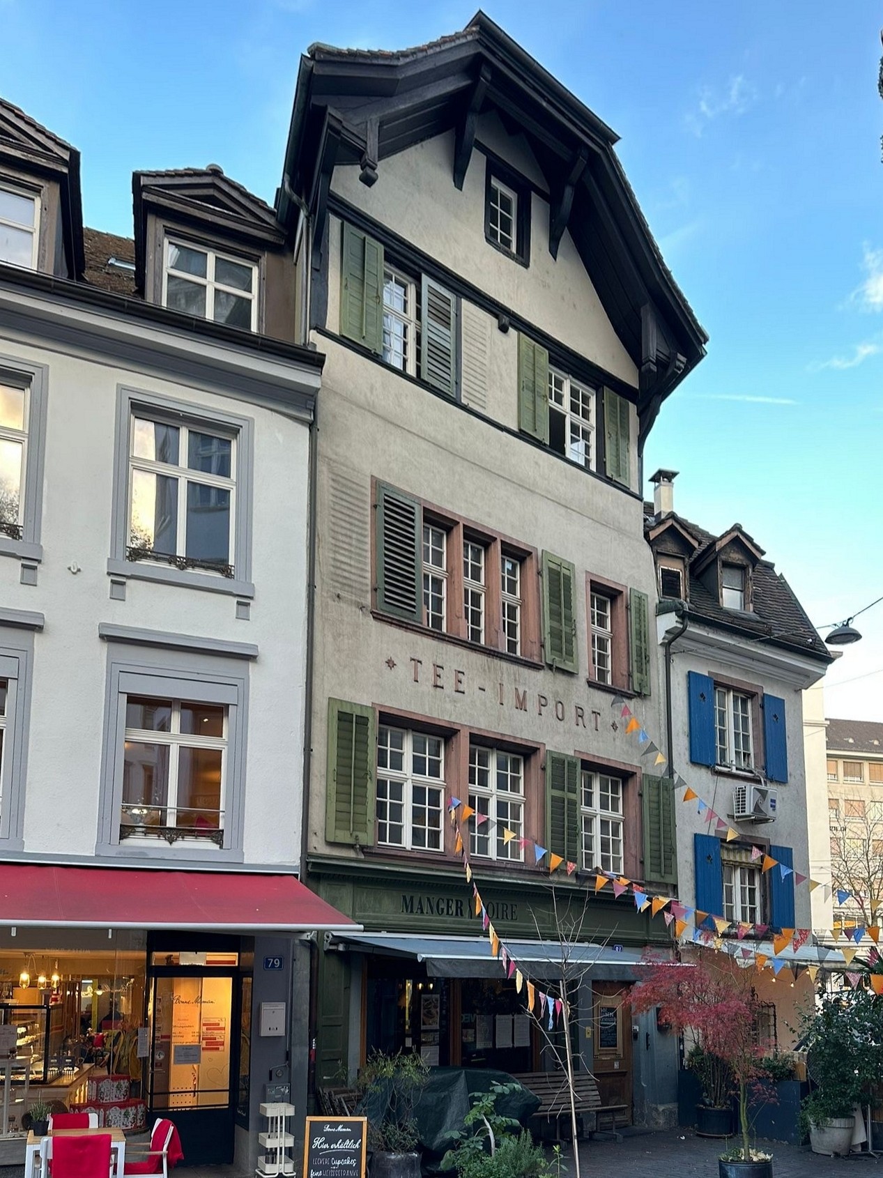 Old building with many windows, different sizes of windows, flags hanging on the building, sign reading 'Tee-Import' and 'Manger la re', awning on the front, potted plants outside