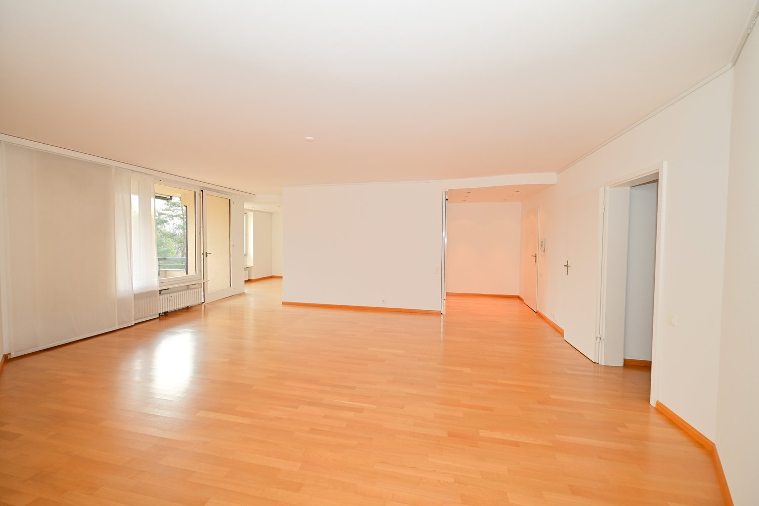 Empty room with wooden floors, white walls, sliding doors, and windows with white curtains