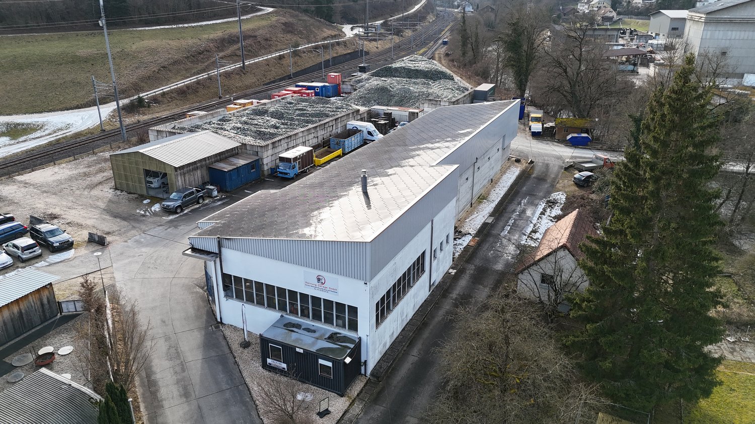 Industrial building with several trucks parked, storage area with containers and a small building.