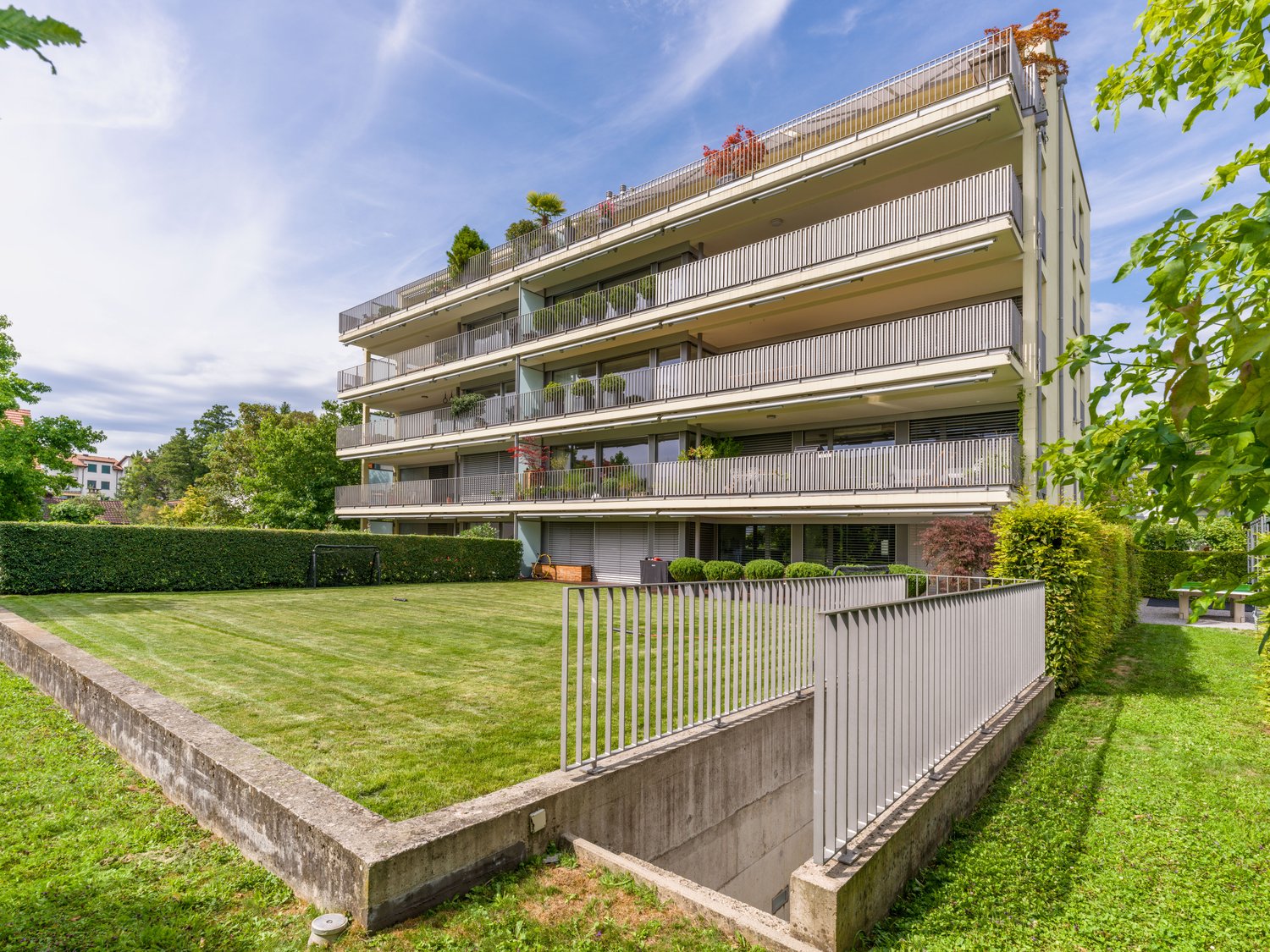 High-rise apartment building with multiple balconies, well-maintained green lawn in front, metal railing along the pathway