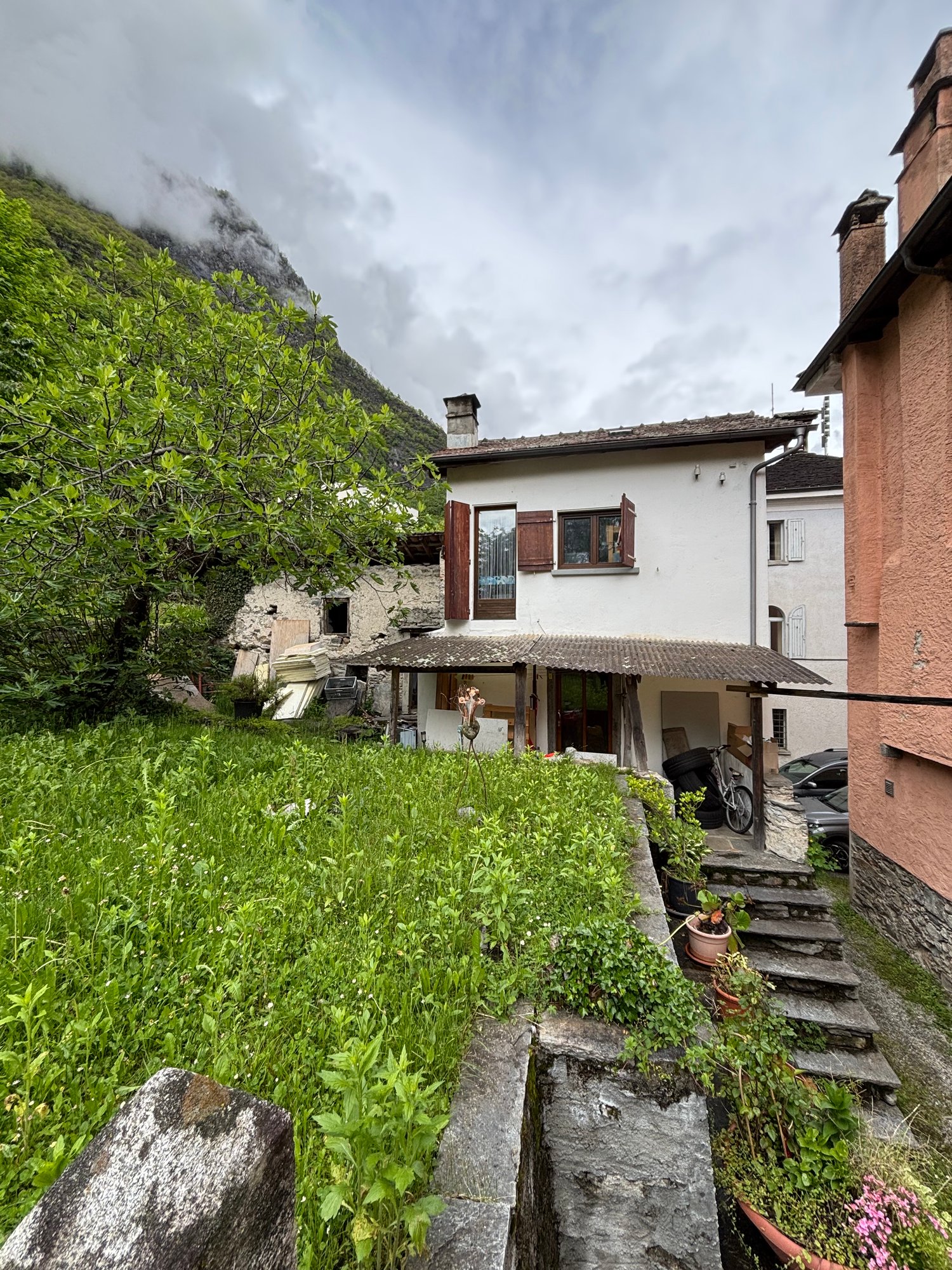 Two-story house with white walls, red roof tiles, and wooden shutters. The house is surrounded by lush greenery, including trees and plants. There is a small balcony or terrace visible on the upper level. The house is situated in a mountainous, rural area