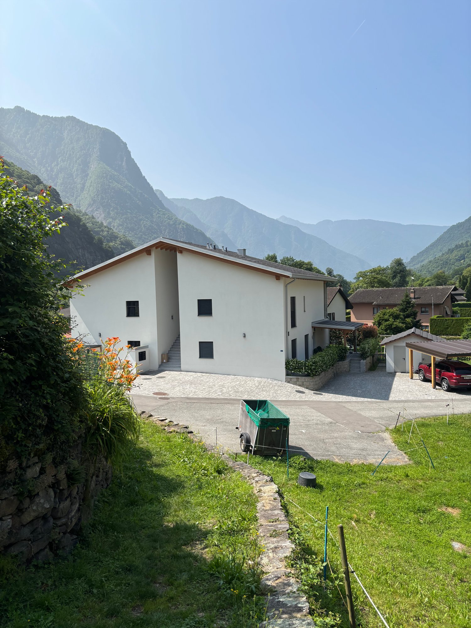 2-story white building with black windows, surrounded by mountains and greenery, with a paved driveway and a green trash bin visible in the foreground