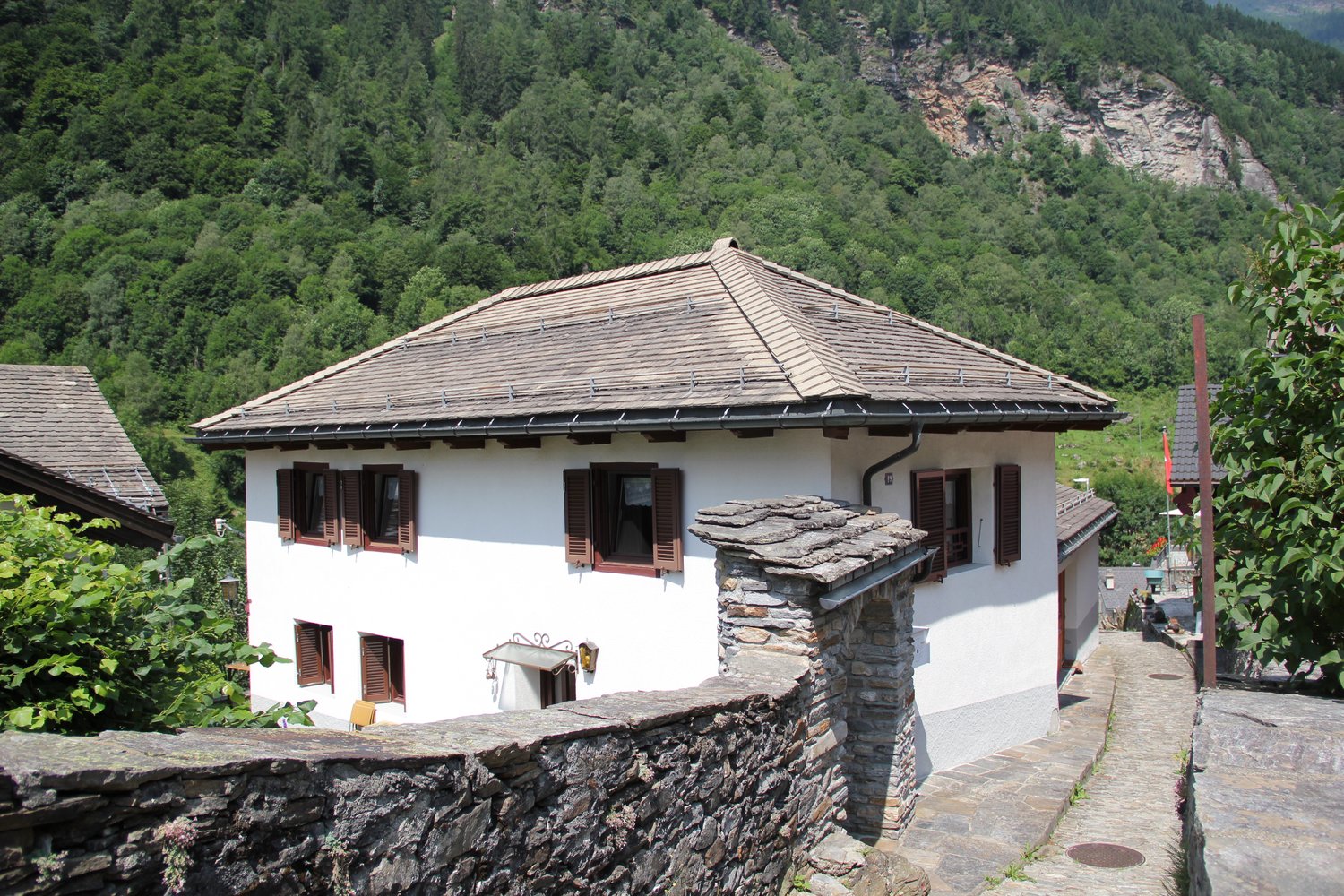 Two-story traditional alpine-style house with a slanted roof, white exterior walls, and wooden shutters. The house is situated on a raised stone foundation, with a stone retaining wall along the front. The surrounding landscape is lush and forested.