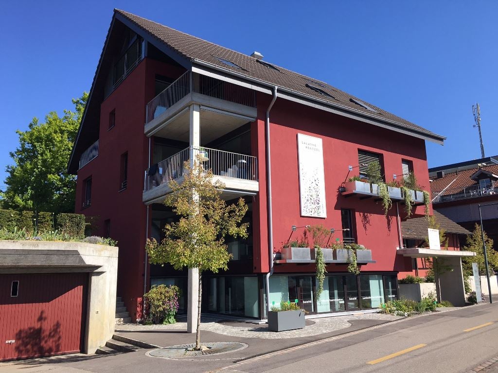 Three-story building, red exterior, balconies, large glass windows, red garage door, greenery, tree, brick paved walkways