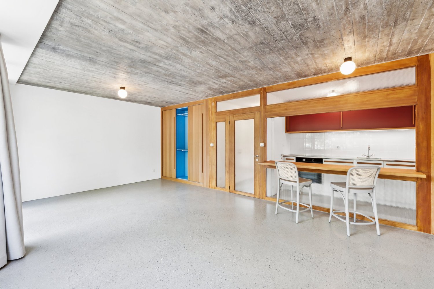 The image shows an open plan living area with a concrete ceiling, wooden walls and frames, and a kitchen area with a counter and stools. The room has a minimalist, modern design with a dishwasher visible in the kitchen area.