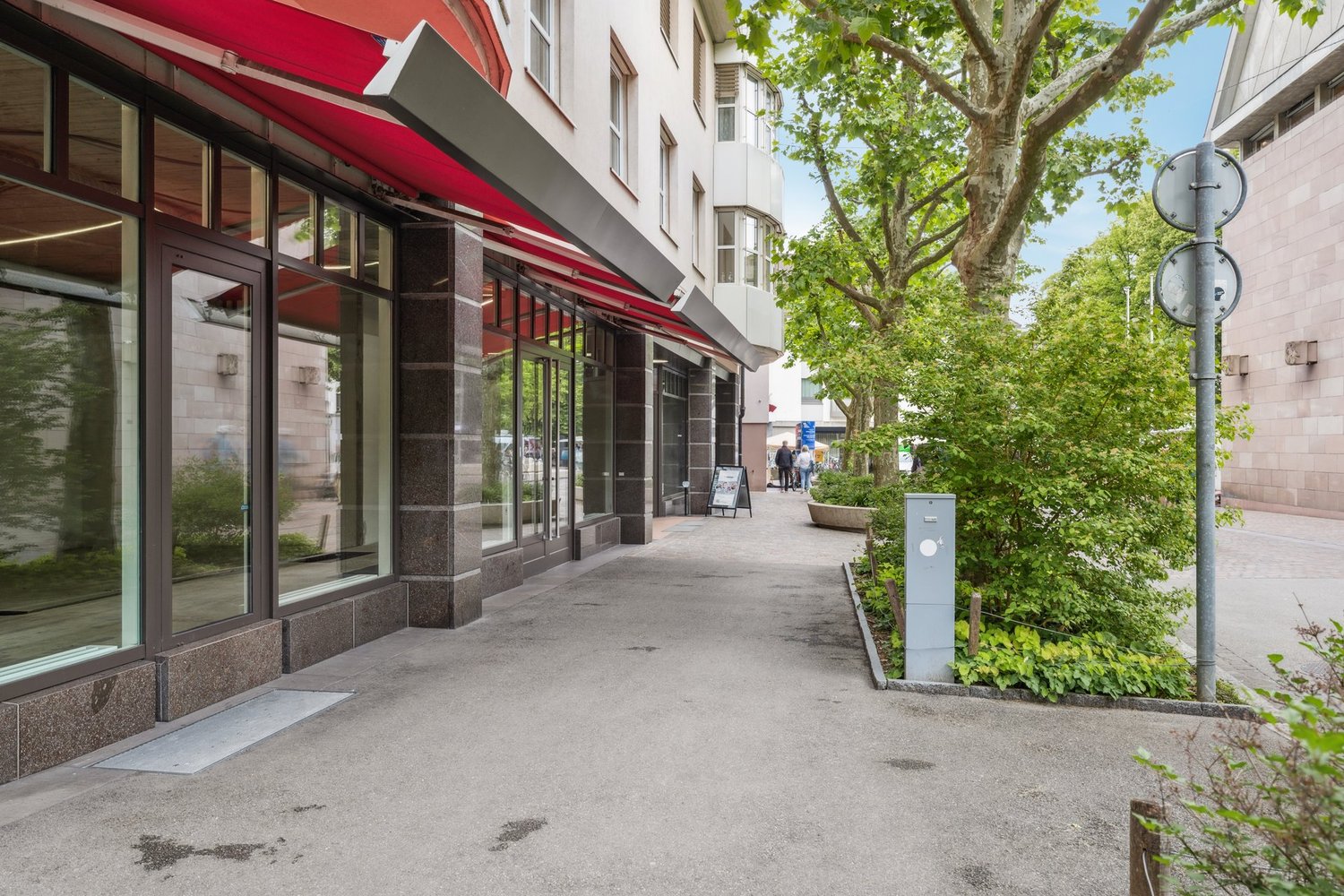 The image shows the exterior of a commercial building with a red awning and large glass windows. The building is surrounded by trees and has a sidewalk in front of it. There are people walking on the sidewalk, and a lamp post is visible.
