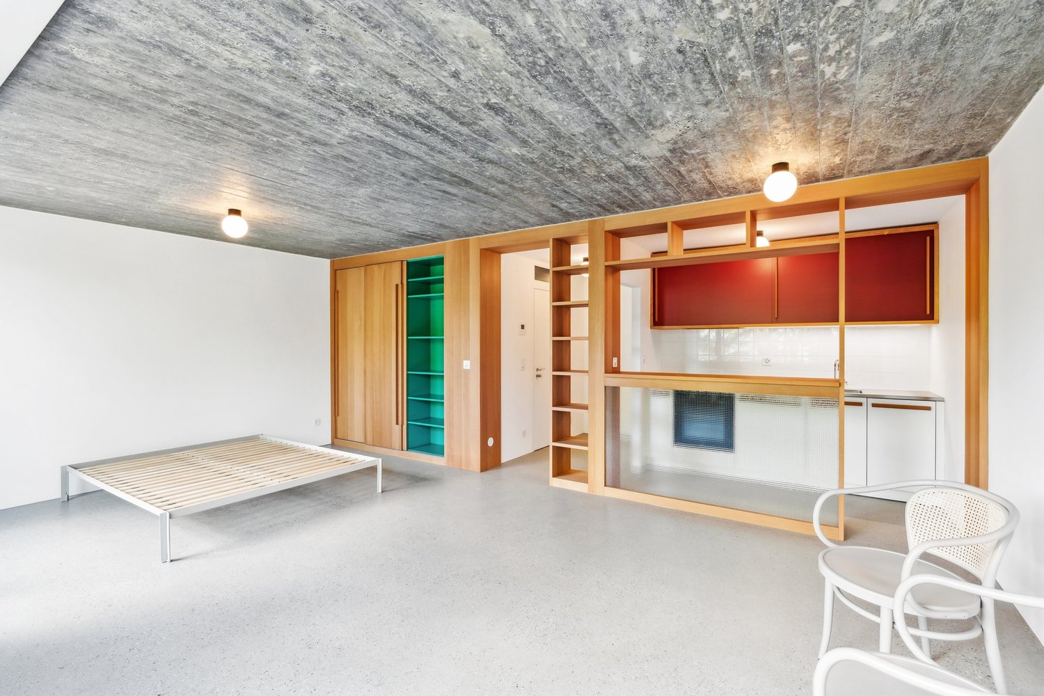 Open-concept studio apartment with concrete ceiling, wooden built-in shelving and cabinets, a bed frame, and a white chair. The room has a minimalist and modern design aesthetic.