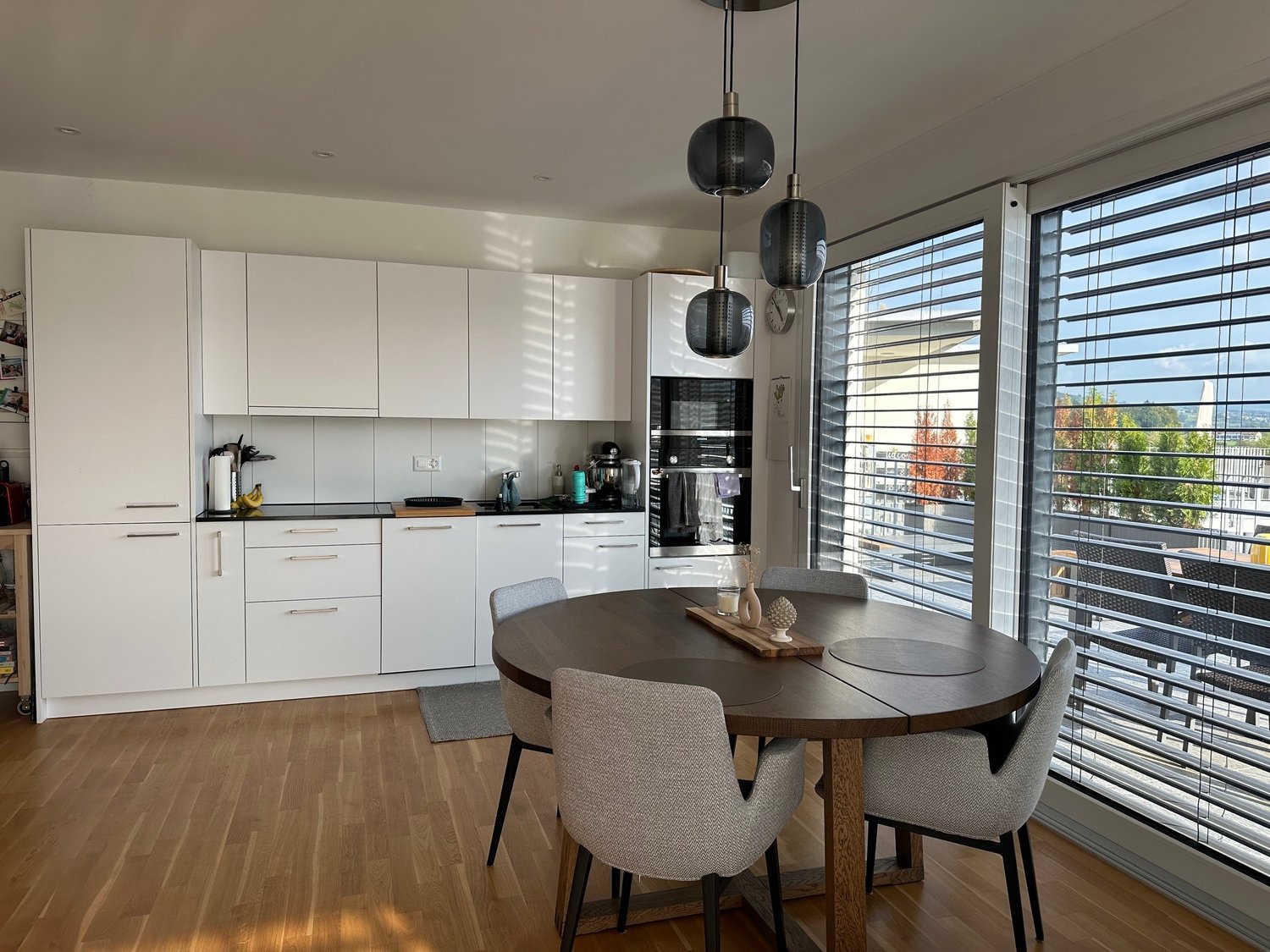 modern kitchen with white cabinets, oak floor, large windows, sliding doors to balcony, round wooden table, grey chairs