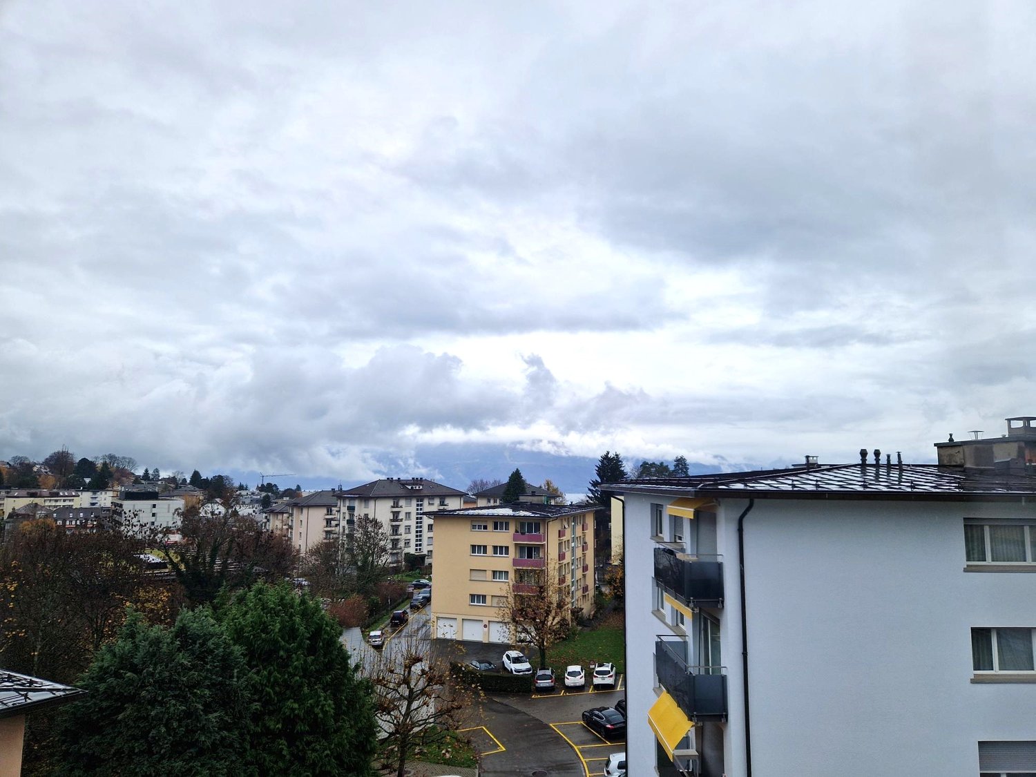 View of an urban area with several multistory buildings, trees, and parked cars. In the distance, there are mountains and a cloudy sky.