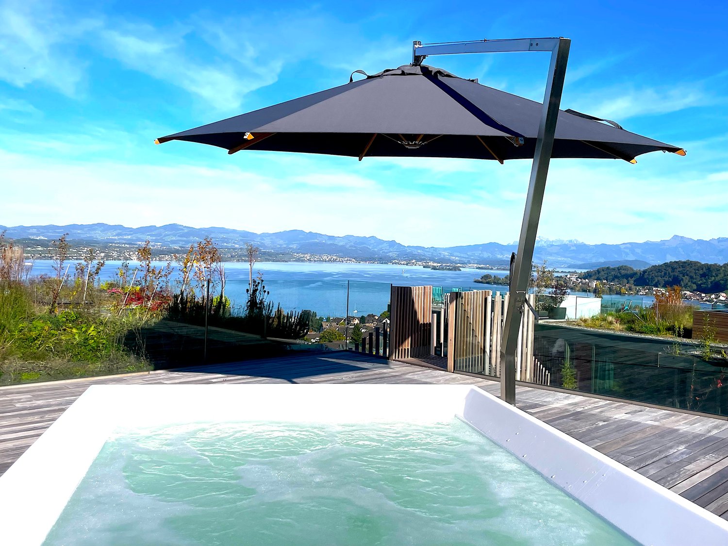 A modern rooftop terrace with a jacuzzi and umbrella overlooking a lake, mountains, and a clear sky.