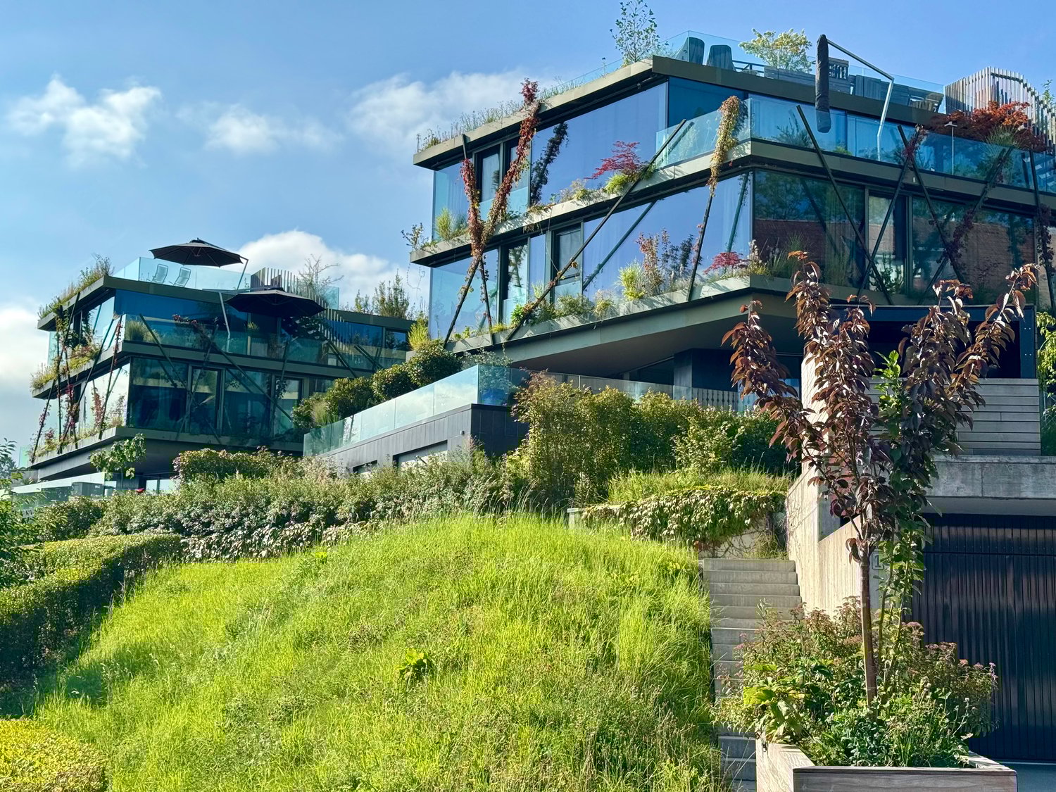 Building on a raised ground, multiple stories, glass walls, greenery on the terrace, outdoor seating, surrounded by vegetation, large garden area, modern design, multiple balconies, staircases leading to the entrance, greenery around the building