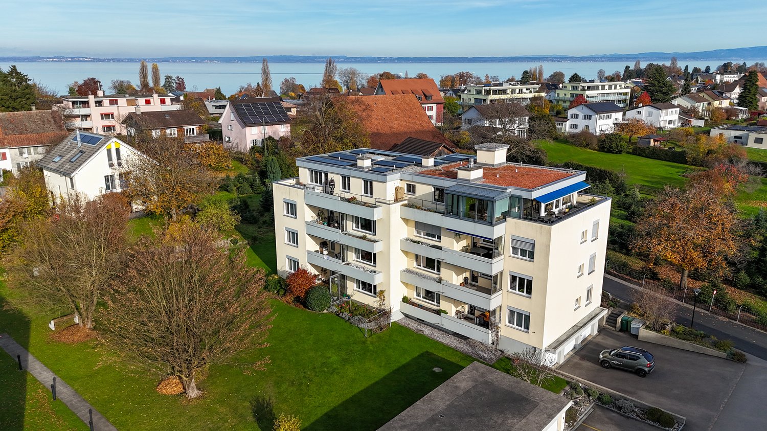 Aerial view of a modern apartment building with multiple balconies, surrounded by landscaped gardens and trees. Nearby buildings, solar panels on the roof, and a lake in the distance. Parking area with a parked car.