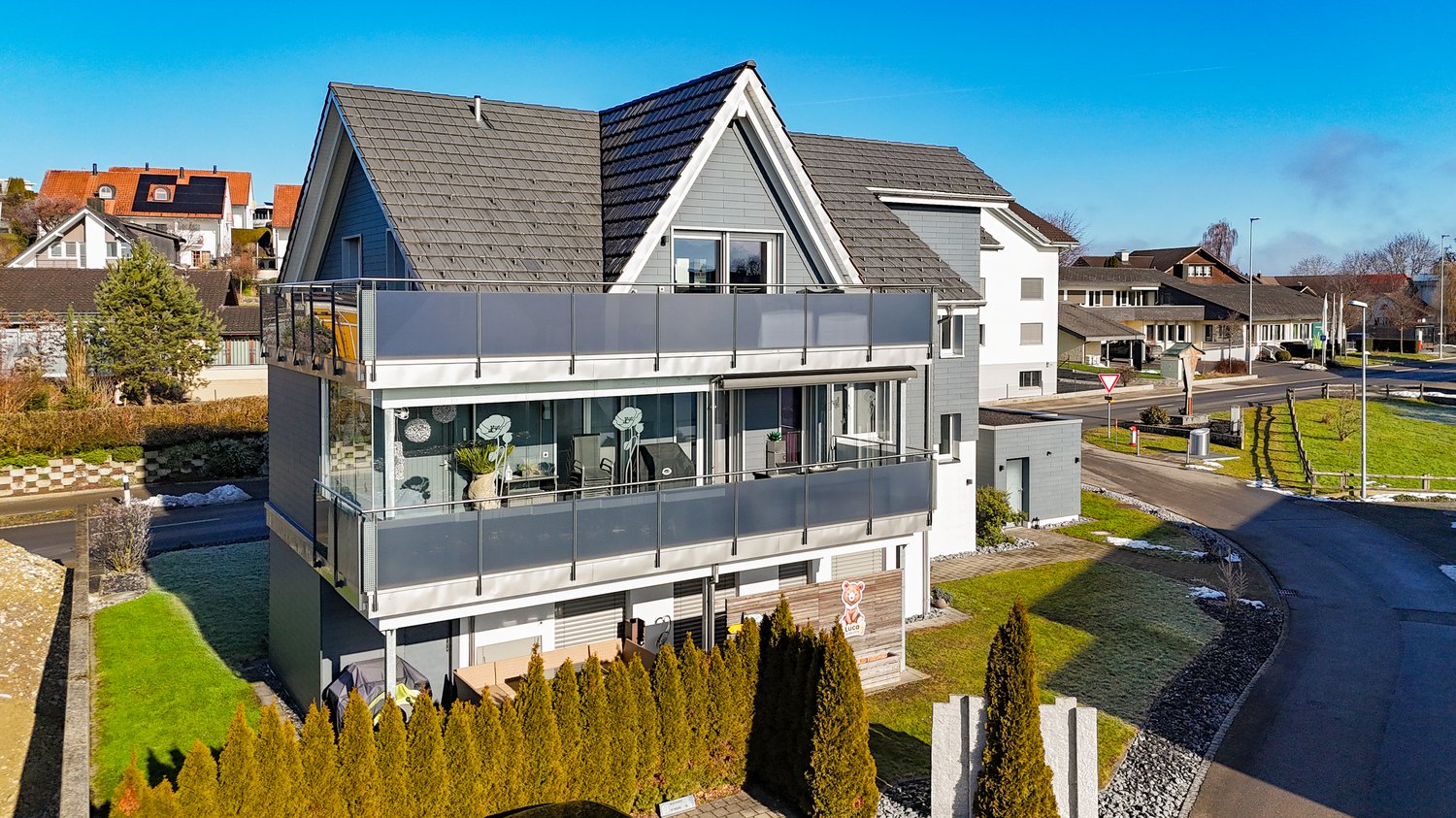 Two-story house, gray exterior, modern design, solar panels on roof, large glass terrace, paved front yard, hedges, nearby road.