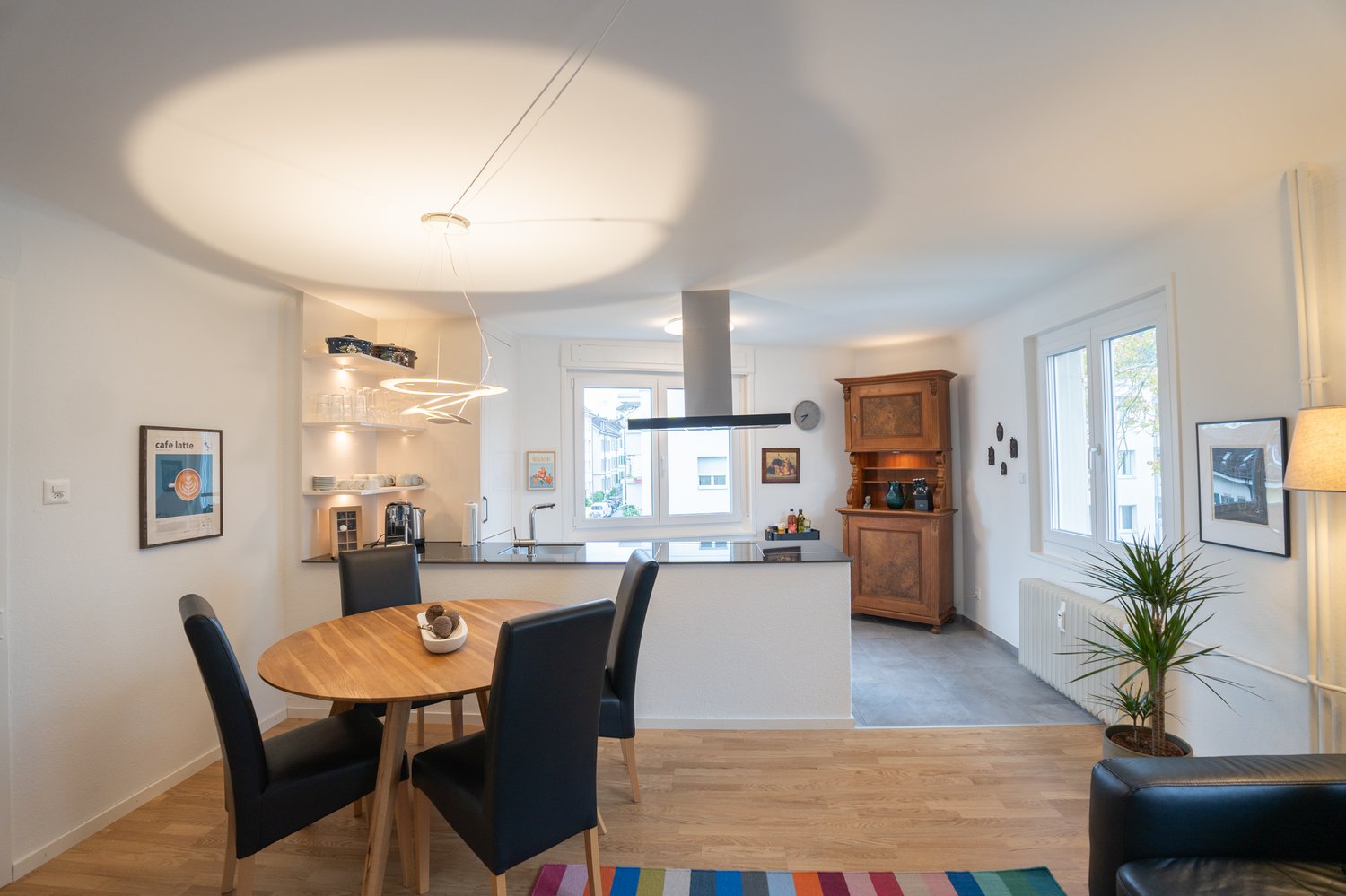 The image shows an open concept living space with a kitchen, dining area, and living room. The kitchen features a black range hood and built-in appliances. The dining area has a round wooden table with black chairs. The living room has a wooden cabinet an