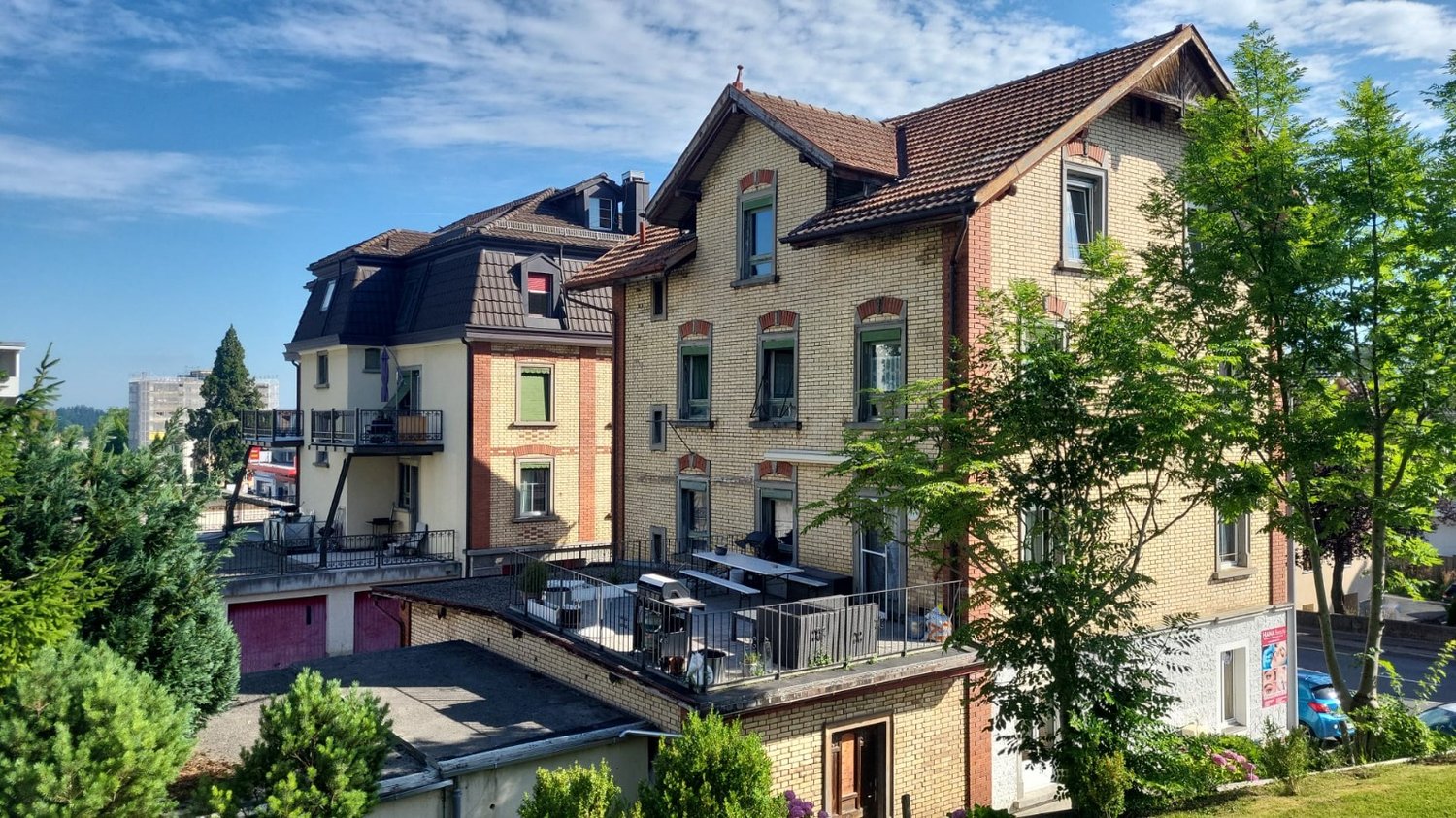 Two story brick house, brown roof, many windows, balcony on the second floor, trees and plants around