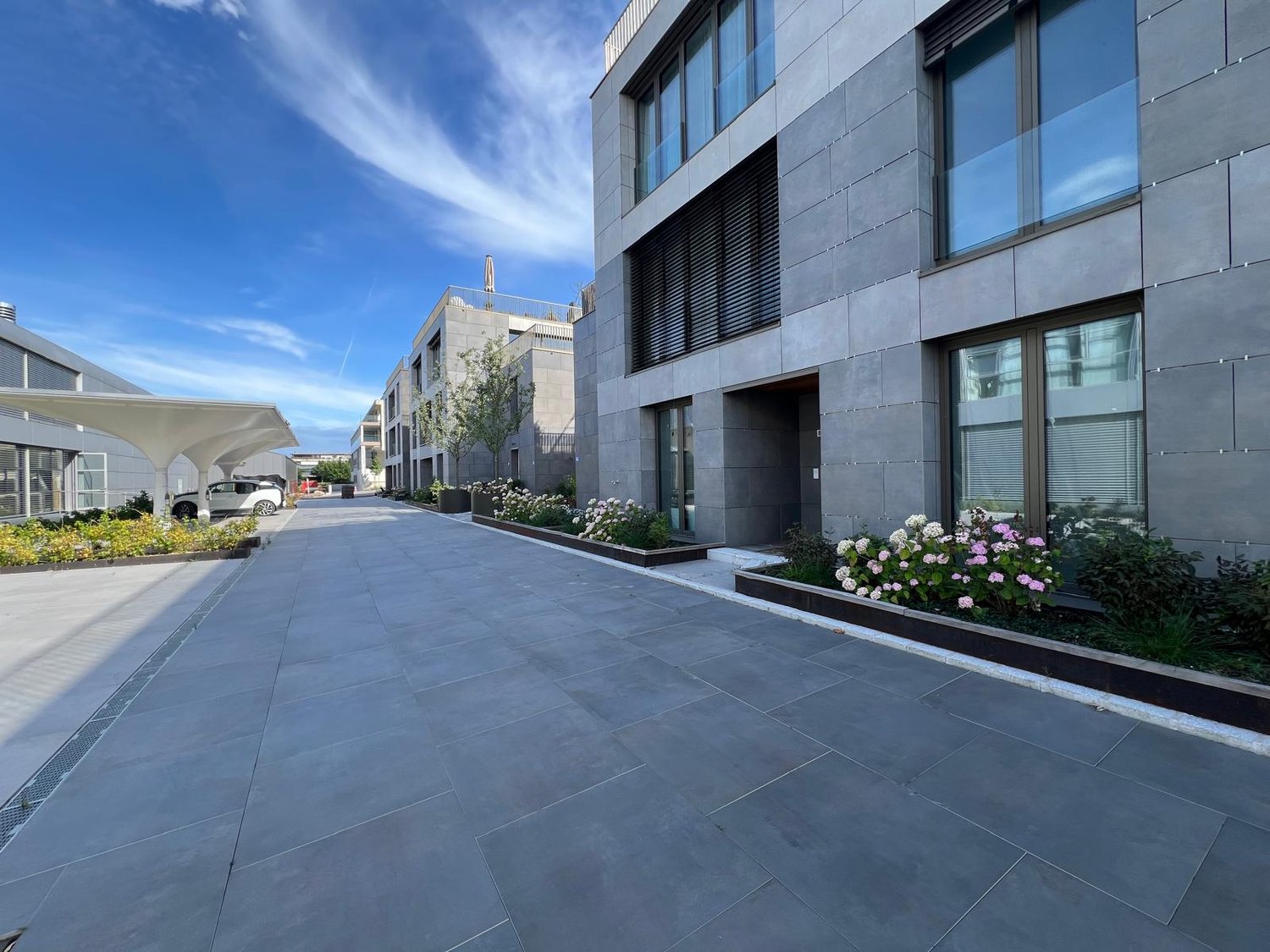Modern multi-story commercial building with glass and concrete facade, paved walkway with landscaping, and a parking area visible in the foreground.