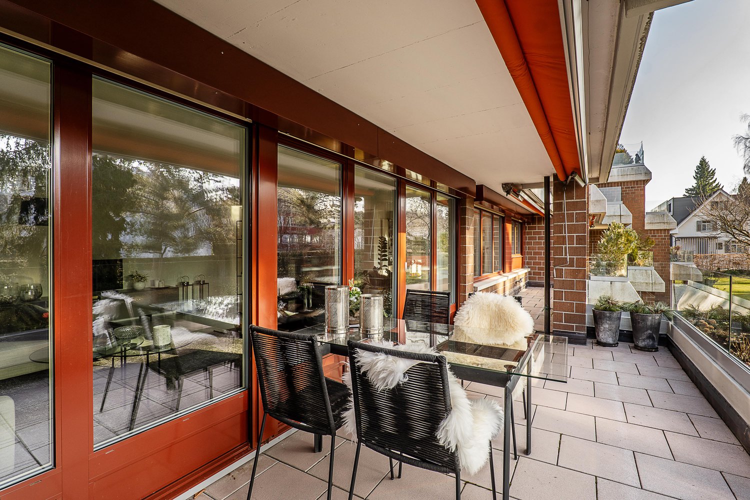outdoor seating area with glass table and chairs, red-framed windows, brick pillar, outdoor plants, metal railings, distant greenery view