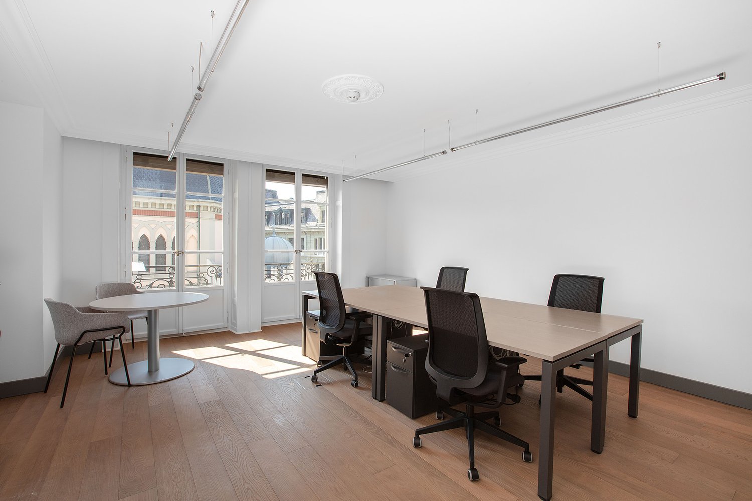 The image shows a modern office space with large windows overlooking a city skyline. The room has a long wooden table with black office chairs, a round table with chairs, and a ceiling with recessed lighting. The floors are hardwood and the walls are whit