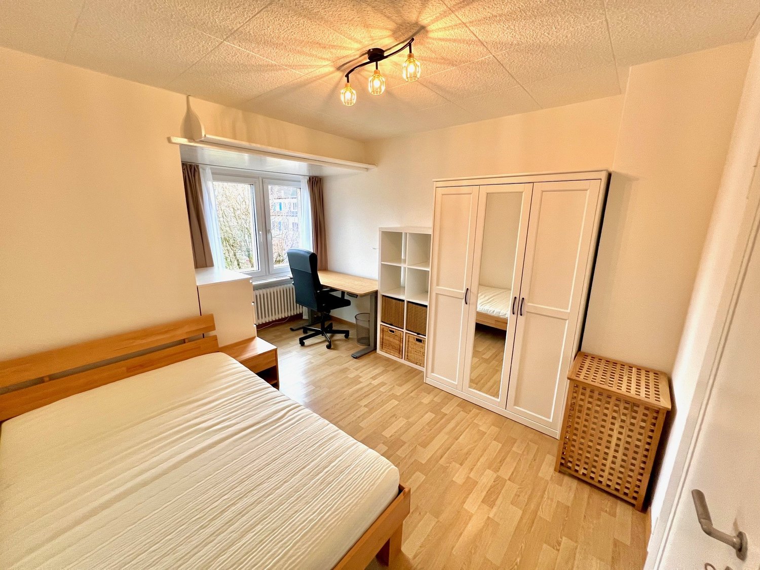 bedroom with a wooden bed, wooden desk, black office chair, white closet, white shelves, radiator, basket, wooden box, white walls, wooden floor, large window with curtains