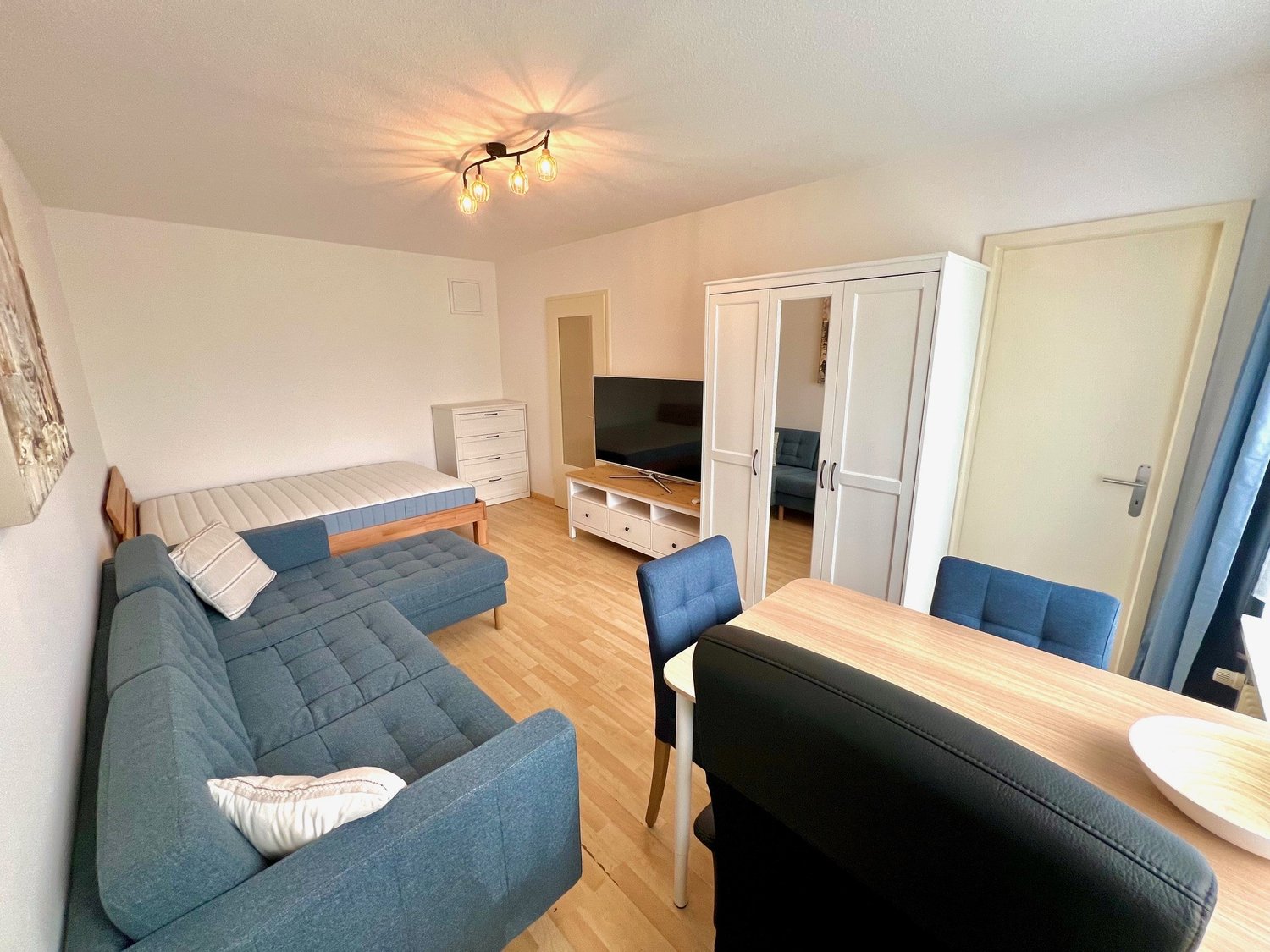 Blue couch, white table, wooden floor, ceiling light, TV, double bed, dresser, wardrobe, blue chair, white bowl on table