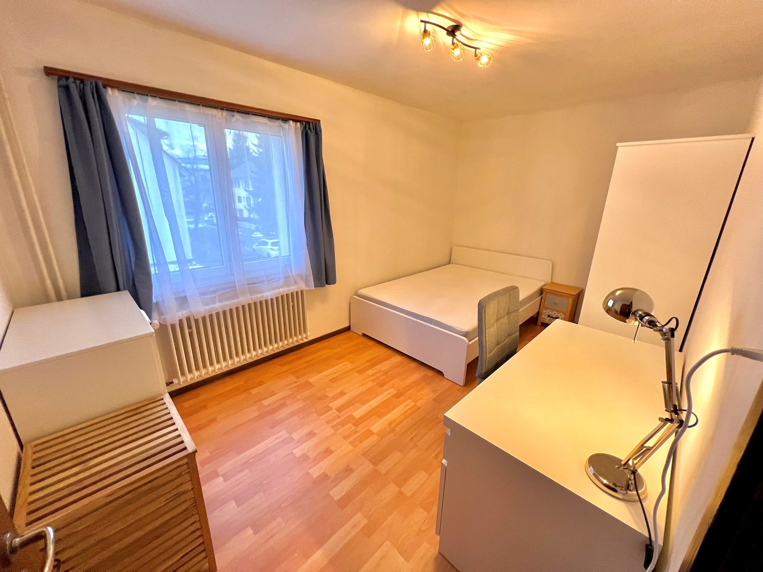 white bed, wooden desk with lamp, white storage units, window with curtains, radiator
