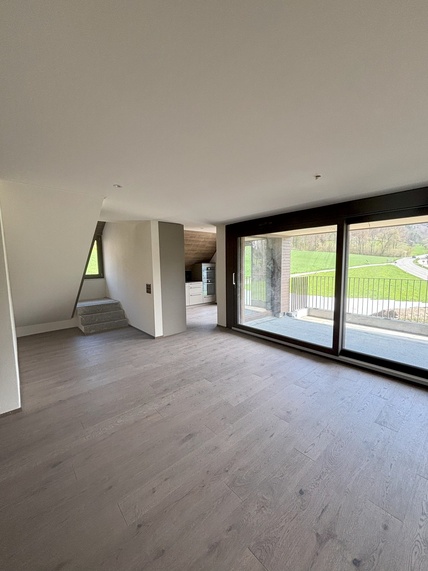 Spacious open-plan living area with large windows overlooking a balcony or terrace, hardwood flooring, and a staircase leading to another level.