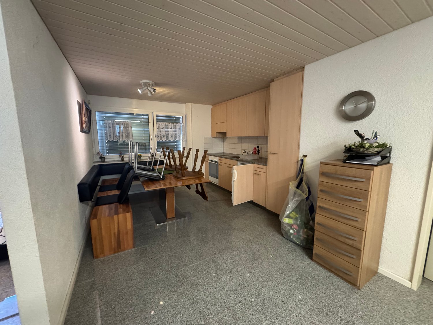 kitchen with dining area, wooden table, wooden bench, wooden cabinets, sink, faucet, wooden floor, white walls, ceiling lamp, window with curtains