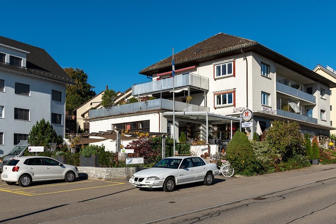 3 story building, white exterior, brown roof, balcony, parking lot, plants, flag